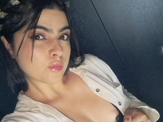 Marilyn Woods con labbra rosa lucide, espressione accigliata, capelli scuri intrecciati, camicia bianca, reggiseno nero visibile, che tiene la camicia, mostrando il décolleté, su sfondo scuro..