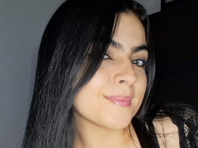 MarilynWoods has black hair, a nose piercing, and a slight smile with glossy pink lips. She's winking one green eye. Her skin is light brown.