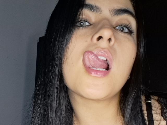 Marilyn Woods, com cabelo preto, olhos verdes e lábios rosados brilhantes, língua para fora, olhando para cima, sobrancelhas escuras e levemente bronzeada, em um selfie, mostrando seu rosto e parte superior do pescoço..