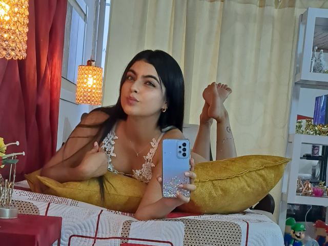 MarilynWoods with long black hair, lying on bed, holding phone, wearing lace top, showing tits, with yellow pillow, crossed legs, looking at camera, red and yellow curtains.