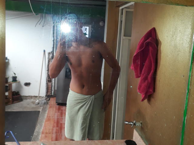 Ein obdachloser Avelio mit einem Handtuch um die Hüfte, der in einem Spiegel-Selfie steht, eine Kamera in der Hand hält, ein rotes Handtuch hängt an einer Tür..