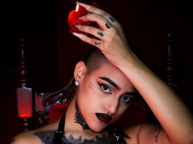 RoseFromHell met een kaalgeschoren hoofd, zwarte lipstick, tatoeages en een neusring, houdt een rode appel vast, er brandt een kaars op de achtergrond, donkerrood, goth, geperforeerde oren, een zwarte choker, een verleidelijke blik..