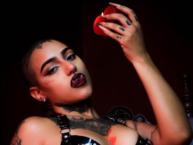 RoseFromHell, com a cabeça raspada, maquiagem escura e tatuagens, segurando um copo de vidro vermelho, língua de fora, usando uma blusa de couro preta, com tatuagens visíveis no peito e piercings, olhando para a câmera..