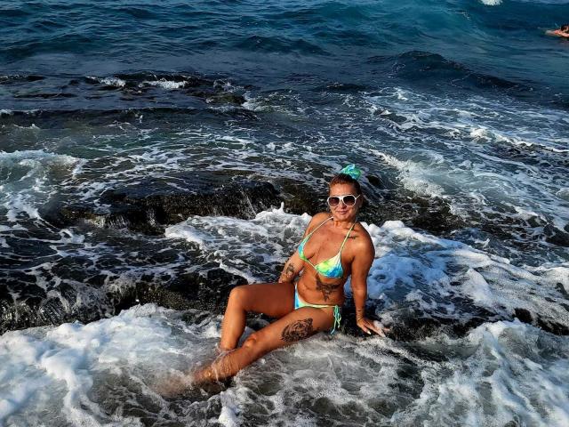 ChaudeEvely, bikini-clad with colorful tits, sitting on dark rocks, waves splashing, tan skin, tattoos on thigh and arm, green hair clip, white sunglasses, ocean background.
