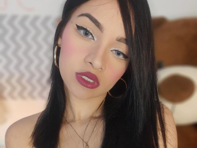 AngelCutie heeft lang zwart haar, roze blush, dikke zwarte eyeliner, rode lipstick en gouden oorringen. Haar huid is licht, en ze kijkt iets opzij..