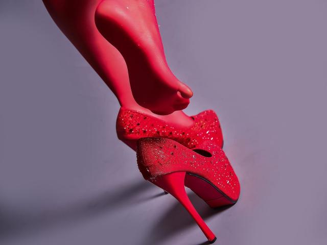 MyraRewe's red stiletto heels with sparkling studs, pointed toes, and tall heels, showing her red-painted legs and foot, standing on a gray surface.