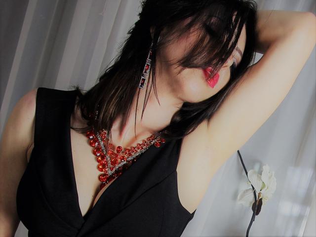 MyraRewe with dark hair, red lipstick, black sleeveless top, and red bead necklace, arm raised, showing her boobs, wearing red earrings, eyes closed, white curtain background.