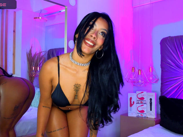 FiorellaBossh with long black hair, smiling, wearing a black bikini top, silver necklace, hoop earrings, tattoos, and a shiny belly button piercing, kneeling on a bed.