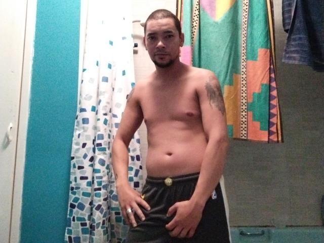 RocioHotMan, shirtless with a trimmed beard, wearing dark pants, has a tattoo on his right shoulder and a slight belly. Standing in a bathroom, looking at the camera.