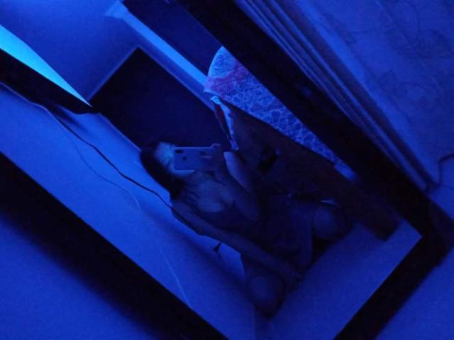 Debora è inginocchiata di fronte a uno specchio, sta scattando un selfie con un telefono, indossa un abito scuro, parzialmente trasparente, con illuminazione di colore blu, con i seni visibili e una tenda sullo sfondo..