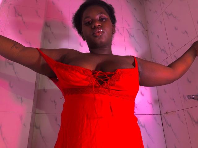 Dilay, dark-skinned, with curly hair, wears a red lace dress, arms out, exposing her boobs, in a pink-lit room, standing against white marble tiles.