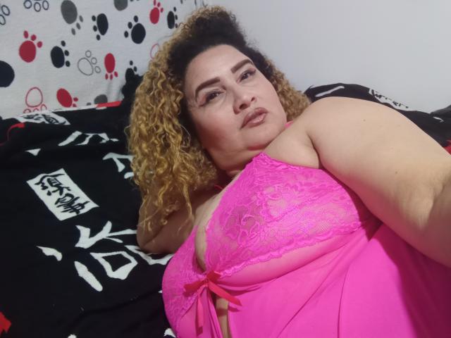 LaPerla69 met krullend blond haar, een roze kanten beha en roze onderbroek, liggend op een zwart-wit bed, kijkend naar de camera met een lichte glimlach. Borsten zichtbaar, zacht licht..