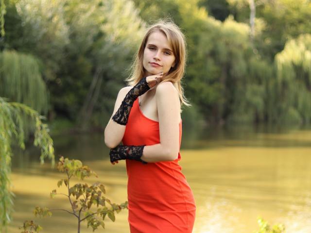 MargaritaKis with light brown hair, wearing a red dress and black lace fingerless gloves, standing by a yellowish lake, touching her chin with a slight smile.