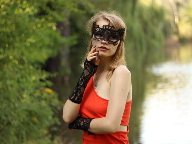 MargaritaKis mit blonden Haaren, die eine schwarze Spitzenmaske, ein rotes Tanktop und schwarze Spitzenhandschuhe trägt, berührt ihr Kinn und steht neben einem grünen, reflektierenden Wasserlauf..