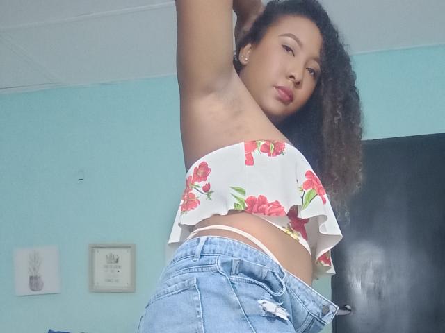 CaroldelaTorre with curly hair, wearing a floral crop top, exposing her small tits, and ripped blue jeans, raising an arm, looking at the camera with a relaxed expression.