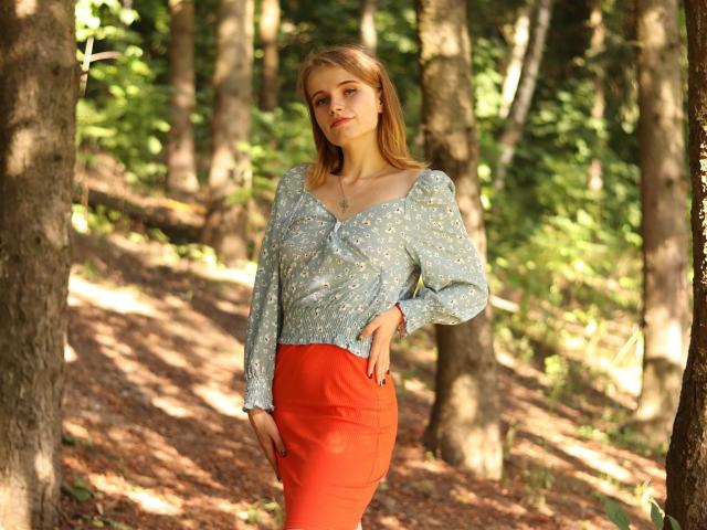 MargaritaKis with blonde hair, wearing a blue floral top and red skirt, stands in a forest, hand on hip, looking slightly to the side, sunlight filtering through trees.