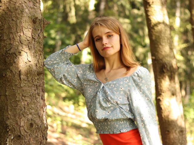 Margarita Kis está de pie en un bosque, con una mano en la cabeza, vistiendo una blusa azul con estampado floral y una falda roja, con una ligera sonrisa, y su trasero ligeramente visible, luciendo segura de sí misma..