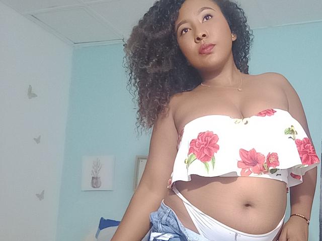 CaroldelaTorre with curly hair, wearing a floral strapless top, showing her tits, and white panties, denim jacket hanging off her shoulder, looking down at the camera.