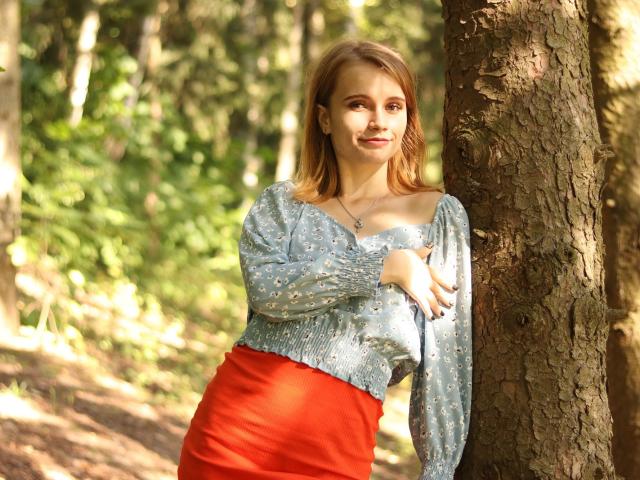 Margarita Kis apoyada en un árbol, vistiendo una blusa azul con lunares y una falda roja, con la mano en el pecho, sonriendo, en un bosque iluminado por el sol..
