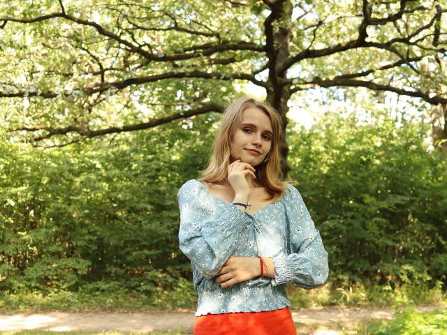 MargaritaKis with blonde hair, light blue floral blouse, and red skirt, standing in a forest, hand on chin, slight smile, green background, sunlight filtering through trees.