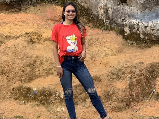 AnnieHal, avec de longs cheveux bruns, portant une chemise rouge avec un motif d'étoile, un jean bleu déchiré et des lunettes de soleil, se tient sur de la terre orange avec un mur de rochers en arrière-plan..