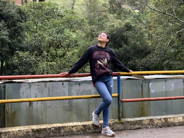 AnnieHal est appuyée contre une rambarde jaune, rouge et bleue, vêtue d'un sweat à capuche noir avec un motif, d'un jean bleu et de baskets blanches, avec ses fesses légèrement relevées et la tête penchée en arrière..