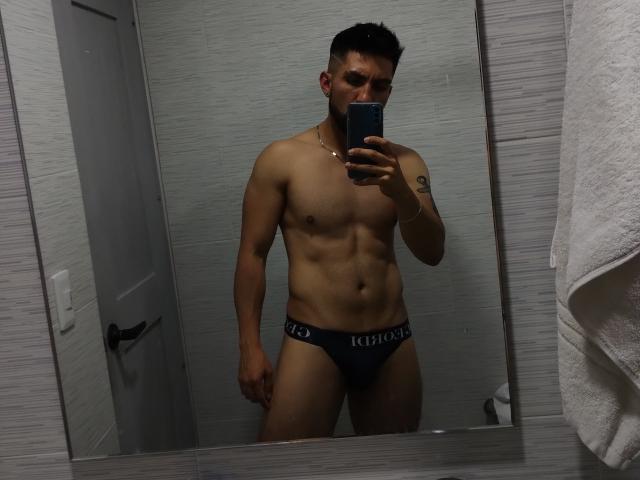 ZacConnor está em frente a um espelho, vestindo uma roupa íntima cinza escura da marca "KISSO", exibindo um corpo musculoso e tonificado, com uma tatuagem visível no braço direito, e um corte de cabelo curto e escuro..