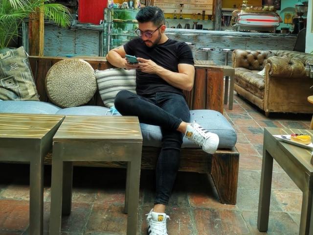 ZacConnor, met een baard en een bril, zit op een houten bank, draagt een zwart T-shirt, versleten jeans en witte sneakers, en typt op een telefoon, omringd door kussens en metalen tafels..