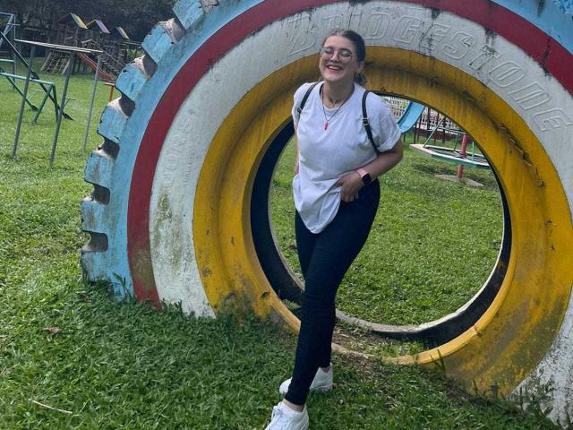 KimSummer è in piedi, sorridente, con gli occhiali, una camicia bianca, pantaloni neri e scarpe da ginnastica bianche, appoggiata a un grande pneumatico circolare e colorato in un parco erboso. Il suo sedere è leggermente visibile..
