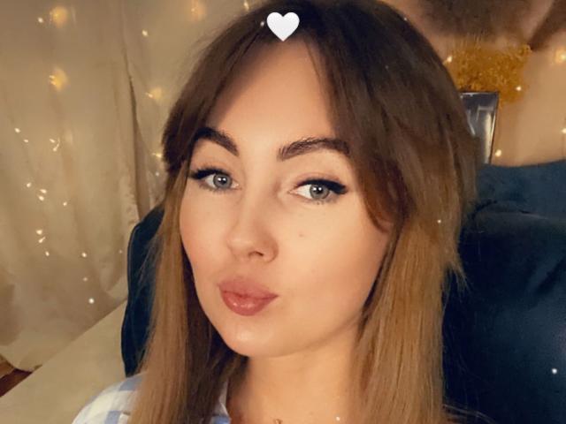 NinaTender with pouty lips, brown hair, blue eyes, and subtle makeup, heart emoji above her head, wearing a white and blue top, soft fairy lights in background.
