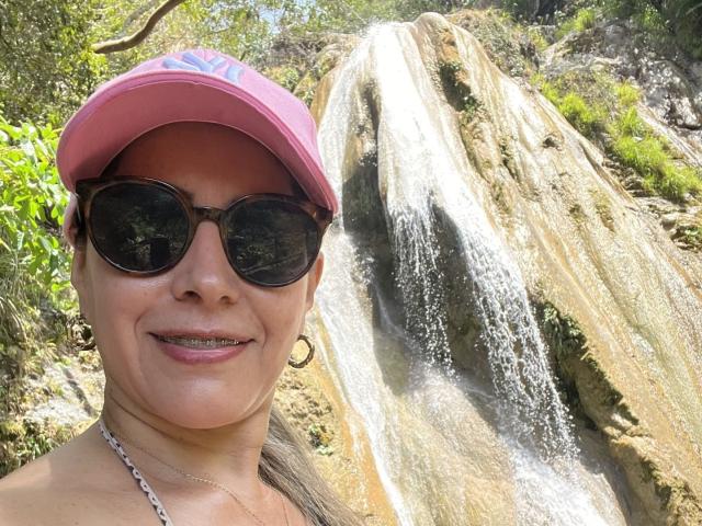 Carolina Ruiz, com boné rosa, óculos de sol escuros, brincos e colar, sorrindo em frente a uma cachoeira, exibindo os seios e o bumbum em uma selfie, usando um colar de corrente..