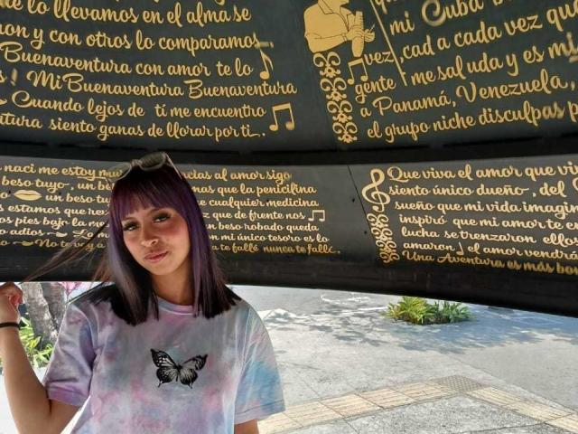 Samantha Morgan, con cabello morado, vistiendo una camiseta con estampado tie-dye y una mariposa negra, de pie frente a una pared negra, sonriendo y sosteniendo un trozo de tela..