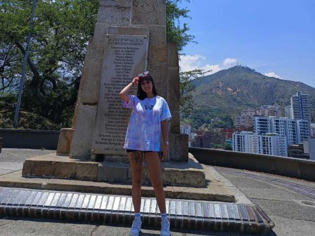 Samantha Morgan está em frente a um monumento de pedra, vestindo uma camiseta estampada, tênis brancos e óculos de sol, com tatuagens na coxa, em um ambiente urbano ensolarado..