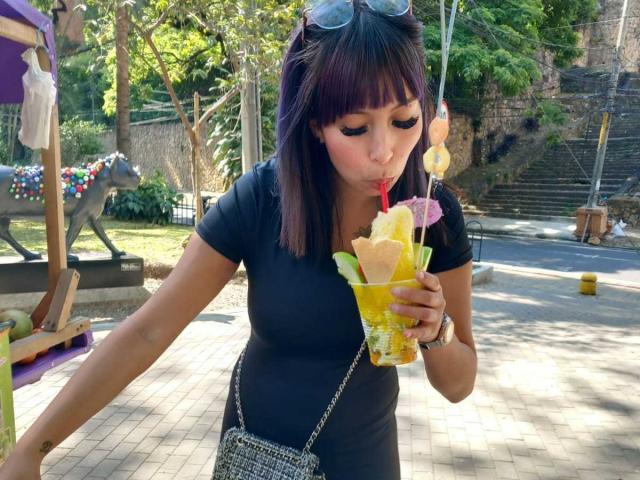 Samantha Morgan, com cabelo roxo escuro, vestido preto e uma pequena bolsa de prata, sugando uma bebida amarela com sorvete e um canudo, em pé ao ar livre, com óculos de sol na cabeça..