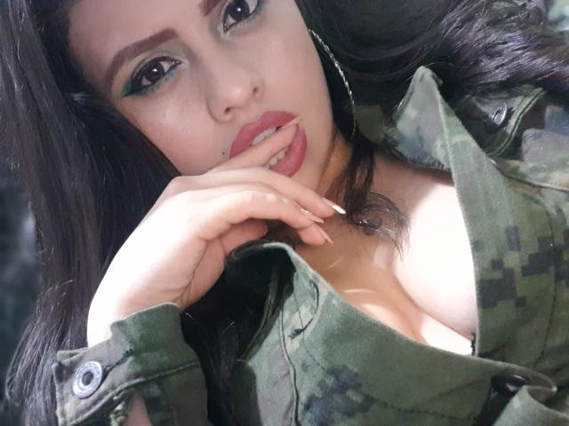 BellaSmithX69 with green eyeshadow, pink lipstick, and hand near her mouth, wearing a camo jacket, exposing her tits, long black hair, and earrings.