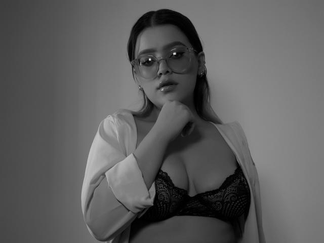 Kim, estate, con occhiali, seno di media grandezza, reggiseno di pizzo nero, camicia bianca aperta, mano vicino alla bocca, grigio in bianco e nero, espressione seria..