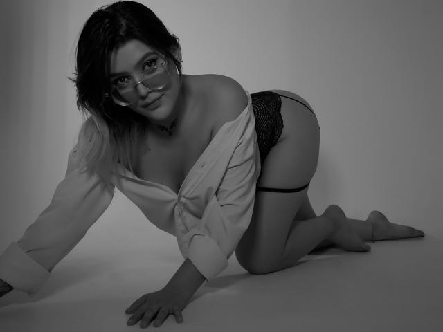 KimSummer à quatre pattes, portant des lunettes, une chemise ouverte et des culottes en dentelle noire, exposant ses seins et ses fesses, avec un tatouage sur son épaule gauche. Photo en noir et blanc..