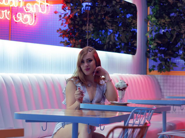 Adela Laurent, com cabelo ruivo e olhos azuis, está sentada em um restaurante retrô, vestindo um vestido listrado com decote baixo, segurando uma colher, com um cupcake na mesa..