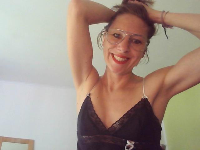 Apolonia, les bras levés, souriante, portant des lunettes et un caraco en dentelle noire, laissant entrevoir ses petites seins et son rouge à lèvres rouge. Ses cheveux bruns sont attachés en un chignon désordonné..