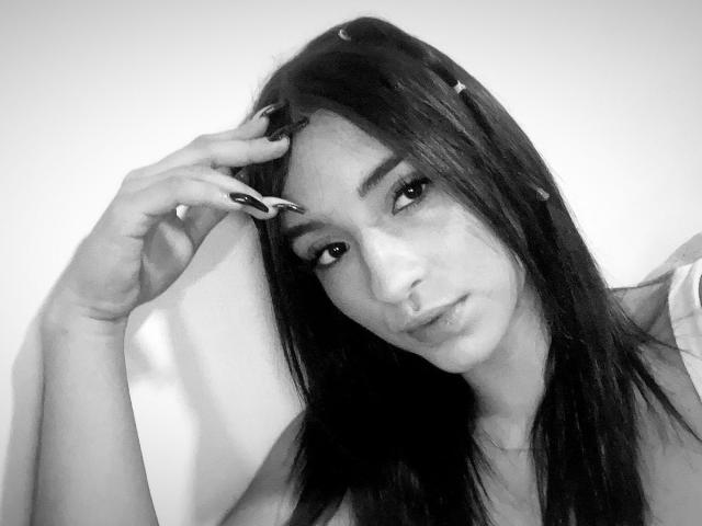 Black-and-white photo of EmmaWaltter with long dark hair, wearing a white shirt, hand touching head, dark nail polish, serious expression, small crescent moon on forehead.