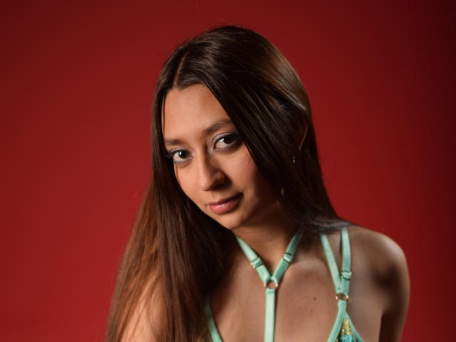 VioletaBlair with long brown hair, green strappy top, small tits, light brown skin, subtle smile, red background, slight head tilt, looking down, soft lighting, gentle expression.