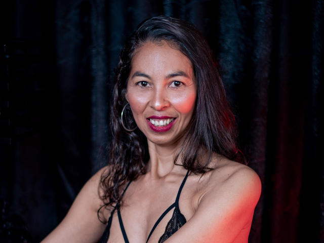 RosalindaCortez with dark wavy hair, smiling, wearing a black lace bra, large hoop earrings, and red lipstick, showing her tits and looking cute.