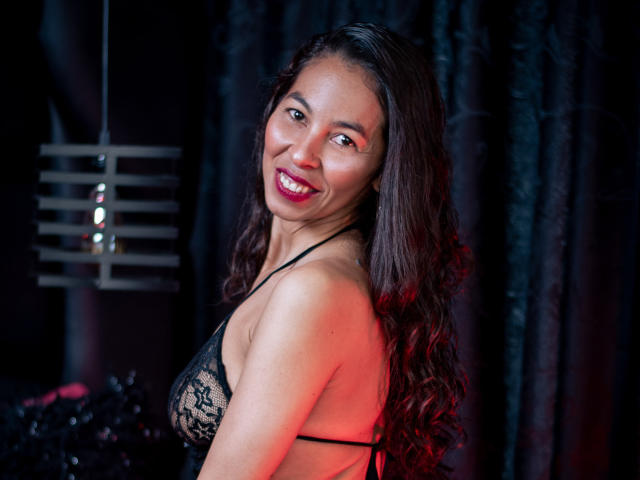RosalindaCortez with long, wavy dark hair, smiling, wearing a black lace bra, showing her small tits, in a dimly lit room with red and black lighting.