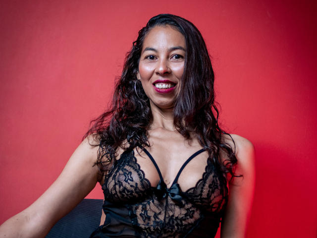 RosalindaCortez with wavy black hair, smiling, wearing a black lace lingerie top with visible tits, against a red background. She's got hoop earrings and a cute bow.