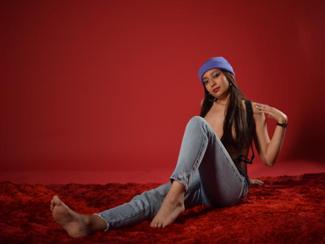 VioletaBlair with long brown hair, blue beanie, bare breasts, and blue jeans, sitting on red furry carpet, barefoot, touching her chin, red background, relaxed, sexy.