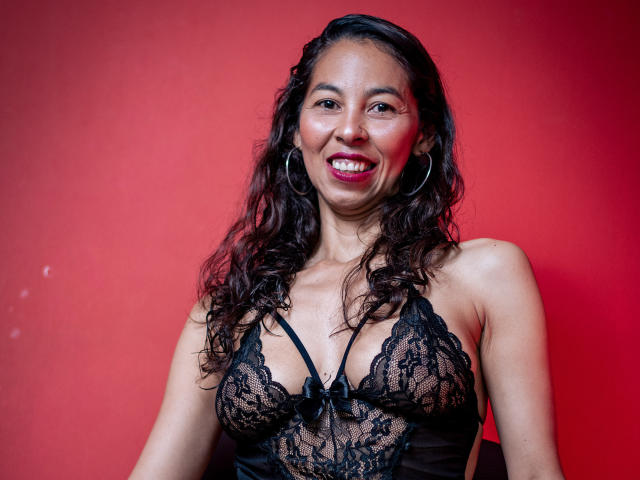 RosalindaCortez with wavy black hair, wearing black lace lingerie, smiling with red lipstick, hoop earrings, and showing her tits against a red background.