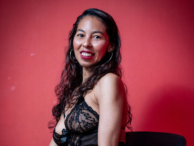 Rosalinda Cortez, met lang, krullend zwart haar, lachend, draagt een zwarte kanten lingerie top, waardoor haar kleine borsten zichtbaar zijn, met grote oorbellen, rode lippenstift, voor een rode muur..