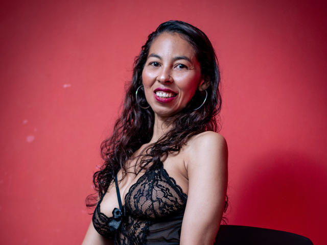 Rosalinda Cortez mit langen, lockigen Haaren, die ein schwarzes Spitzen-BH-Oberteil trägt, lächelt mit rotem Lippenstift, trägt Creolen und hat ein sichtbares Dekolleté, vor einem roten Hintergrund..