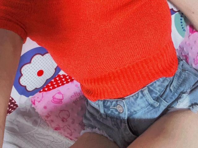AmberGray69 trägt ein hellorangefarbenes Rippstrickoberteil und zerrissene blaue Jeansshorts, die ihre schlanke Taille und die oberen Oberschenkel zeigen. Ihr Oberteil ist eng geschnitten und betont ihre kleinen Brüste..