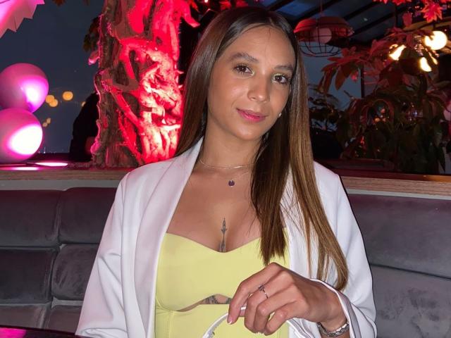 SarahWest, con lunghi capelli castani, rossetto rosa, giacca bianca, top giallo, seno piccolo, che tiene una borsa bianca, con un anello al dito, un tatuaggio sotto il décolleté, che guarda la fotocamera, illuminazione rossa..